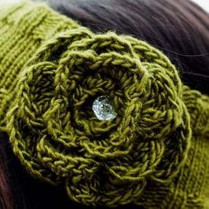 May include: Close-up of an olive green knitted headband featuring a large, textured flower with a sparkling crystal centre. The headband is made of thick yarn and has a textured pattern. The word "Pattern" is visible.