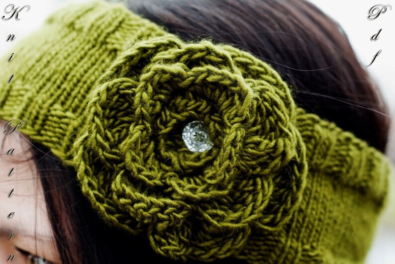 May include: Close-up of an olive green knitted headband featuring a large, textured flower with a sparkling crystal centre. The headband is made of thick yarn and has a textured pattern. The word "Pattern" is visible.