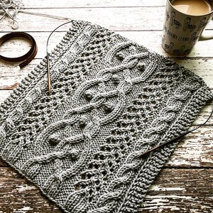May include: A gray knitted blanket with a cable knit pattern. The blanket is lying on a wooden surface with a cup of coffee and a knitting needle.