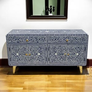 以下が含まれることがあります： 白のフローラルパターンが施されたネイビーブルーのキャビネット。キャビネットには、4つのドアと2つの引き出しがあり、金色の金具が付いています。キャビネットは金色の脚で支えられています。家具です。