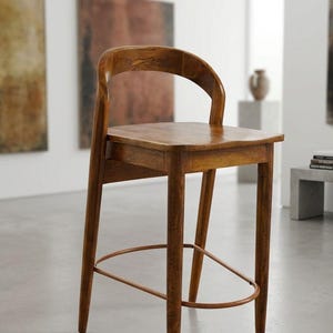 May include: A wooden bar stool with a curved backrest and a square seat. The stool has four tapered legs and a circular footrest. The wood has a warm, medium brown tone. The stool is in a room with artwork on the walls.