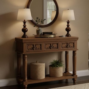May include: A dark wood console table with elephant carvings on the drawers, topped with two lamps with white shades and a round mirror. A lower shelf features woven baskets and a plant pot. The table is a stylish piece of furniture for a hallway or living room.
