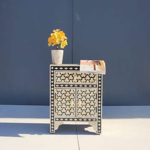 May include: A white and black patterned bedside table with a drawer and two doors. The table has a geometric design and is topped with a small vase of yellow flowers and a magazine. The table is 61 cm tall.