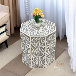 Könnte beinhalten: Ein grauer, sechseckiger Beistelltisch mit weißem Blättermuster. Der Tisch hat eine kleine weiße Vase mit gelben Blumen. Der Tisch ist etwa 51 cm hoch.