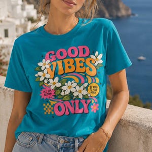 Può includere: T-shirt turchese con le parole "GOOD VIBES ONLY" in rosa, arancione e bianco, con accenti floreali. La maglietta ha anche le frasi "LET JOY BLOOM" e "BE KIND TO YOUR MIND". La persona indossa jeans blu.