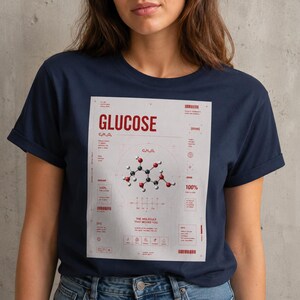 Può includere: T-shirt blu navy con grafica di una molecola di glucosio e la parola "GLUCOSE" in rosso. Il design include la formula chimica C6H12O6 e altri dettagli scientifici. La maglietta è realizzata in un materiale morbido.