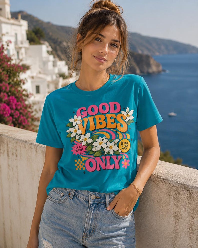 Può includere: T-shirt turchese con le parole "GOOD VIBES ONLY" in rosa, arancione e bianco, con accenti floreali. La maglietta ha anche le frasi "LET JOY BLOOM" e "BE KIND TO YOUR MIND". La persona indossa jeans blu.