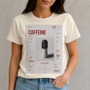 Può includere: T-shirt color crema con un design grafico rosso. Il design presenta la parola "CAFFEINE", un'illustrazione di capsula e dettagli scientifici. Include anche il testo "FUEL YOUR MIND, MASTER YOUR DAY".