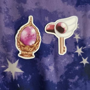 Puede incluir: Dos pegatinas con artículos de transformación de chicas mágicas. Una es una varita dorada y rosa en forma de huevo con una gema rosa. La otra es una llave dorada con una cabeza de pájaro blanca y un solo ojo.