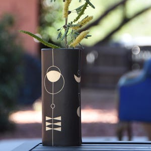 May include: A black cylindrical vase with a geometric design in white, featuring circles, lines, and crescent shapes. The vase holds a sprig of greenery with yellow flowers. The vase is approximately 8 inches tall.