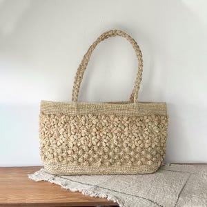 Op de afbeelding: Een beige geweven draagtas met een bloemenpatroon. De tas heeft twee gevlochten handvatten en een rechthoekige vorm. De body van de tas is bedekt met kleine, verhoogde bloemvormen. De tas staat op een houten oppervlak.