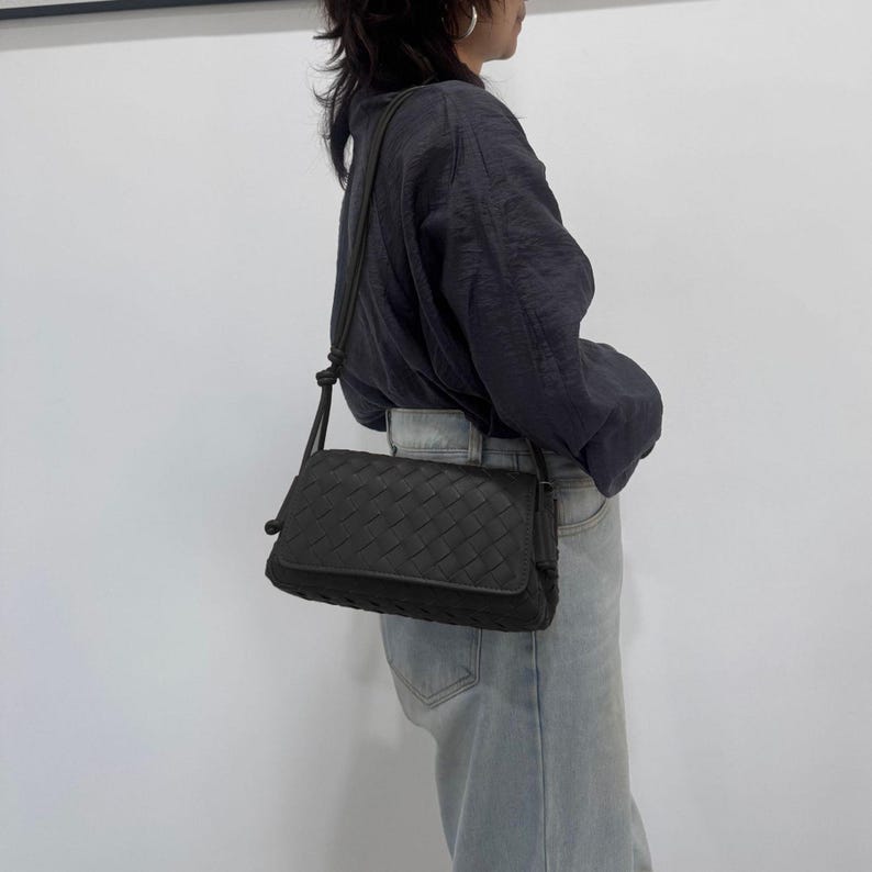 Op de afbeelding: Een zwarte crossbody tas met een geweven patroon. De tas heeft een rechthoekige vorm met een klepsluiting en een lange, verstelbare riem. De tas is gemaakt van donker materiaal en wordt over de schouder gedragen.