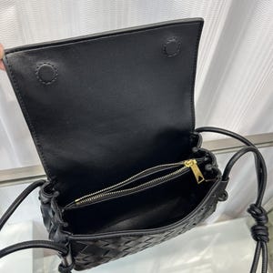 Op de afbeelding: Zwarte leren crossbodytas met een geweven design aan de voorkant. De tas heeft een klepsluiting met twee drukknopen en een gouden ritssluiting. De riem is een dun, verstelbaar leren koord.
