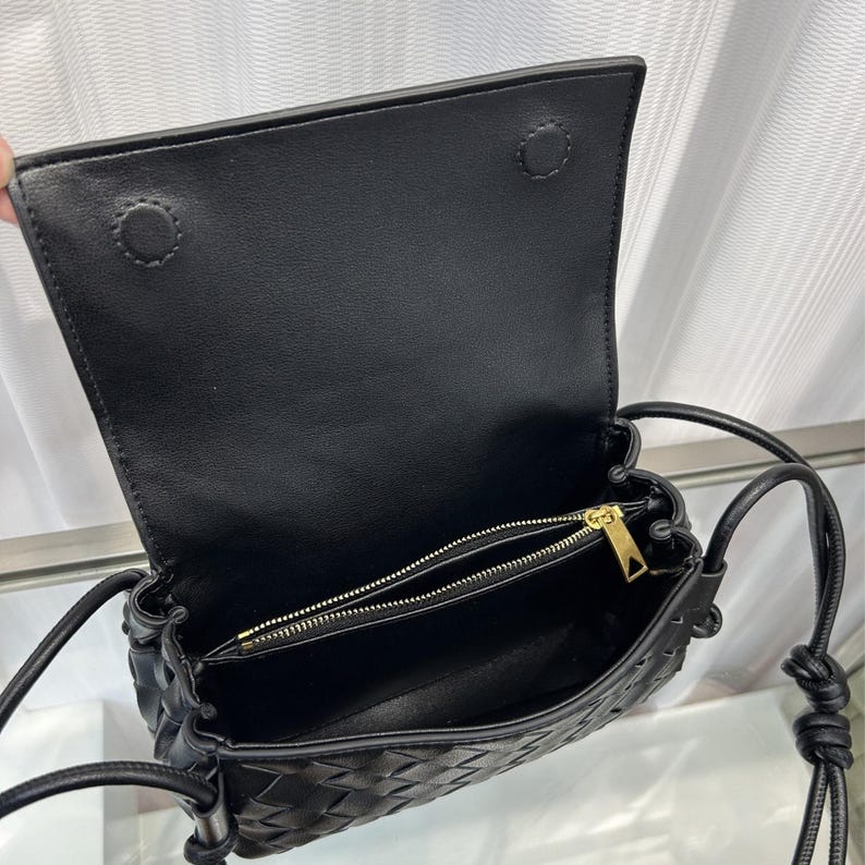 Op de afbeelding: Zwarte leren crossbodytas met een geweven design aan de voorkant. De tas heeft een klepsluiting met twee drukknopen en een gouden ritssluiting. De riem is een dun, verstelbaar leren koord.
