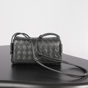 Op de afbeelding: Zwarte leren crossbody tas met een geweven ruitpatroon en geknoopte details. De tas heeft een rechthoekige vorm en een lange, dunne riem. De tas staat op een zwarte ondergrond.