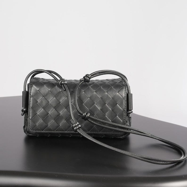 Op de afbeelding: Zwarte leren crossbody tas met een geweven ruitpatroon en geknoopte details. De tas heeft een rechthoekige vorm en een lange, dunne riem. De tas staat op een zwarte ondergrond.