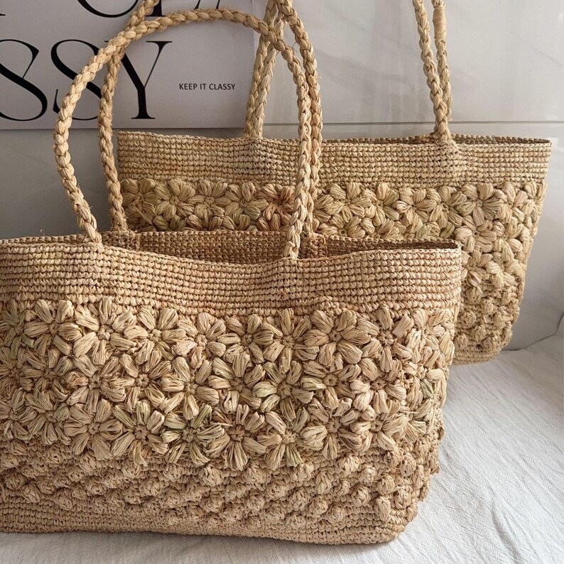 Op de afbeelding: Twee geweven strooien draagtassen met bloemendetails. De tassen hebben gevlochten handvatten en zijn in een natuurlijke beige kleur. De tassen zijn versierd met een herhalend patroon van geweven bloemen.