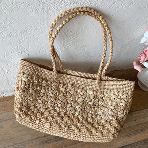 Op de afbeelding: Een beige geweven strooien tote bag met een bloemenpatroon. De tas heeft gevlochten handvatten en een rechthoekige vorm. De tas staat op een houten oppervlak.