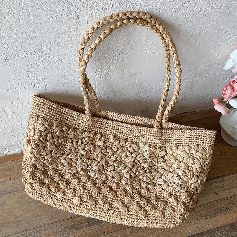 Op de afbeelding: Een beige geweven strooien tote bag met een bloemenpatroon. De tas heeft gevlochten handvatten en een rechthoekige vorm. De tas staat op een houten oppervlak.