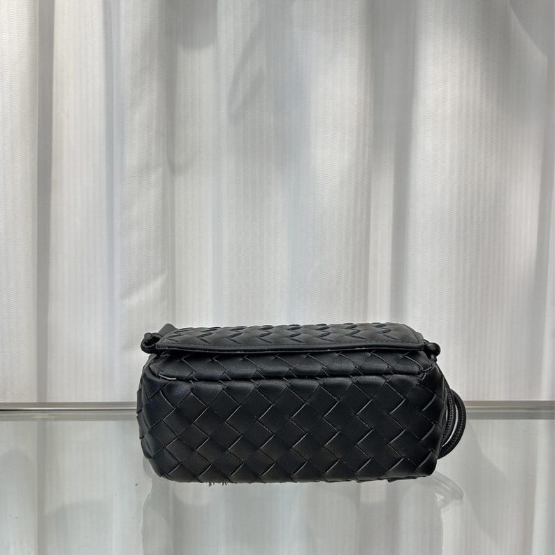 Op de afbeelding: Zwarte crossbodytas van leer met een geweven patroon. De tas heeft een rechthoekige vorm en een klepsluiting. Het geweven patroon bedekt de hele tas en heeft een dunne, verstelbare riem. Ongeveer 20 cm lang.