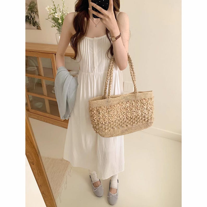 Op de afbeelding: Een witte zomerjurk met spaghettibandjes en een geweven strooien tote bag. De tas is versierd met kleine, ronde, beige versieringen. De outfit wordt gecombineerd met grijze ballerina's. Een lichtblauw vest wordt in de arm gehouden.