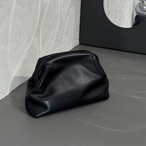 May include: A black, gathered clutch bag made of smooth material. The bag is rectangular with rounded edges and a soft, voluminous shape. It sits on a gray surface, with a textured white wall in the background.