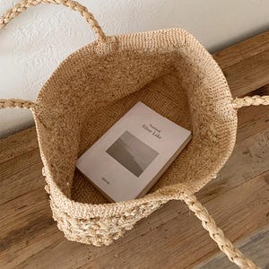 Op de afbeelding: Een beige geweven strooien draagtas met gevlochten handvatten, op een houten oppervlak. In de tas ligt een boek met de titel "Notebook Silver Lake". De tas heeft een getextureerde uitstraling.