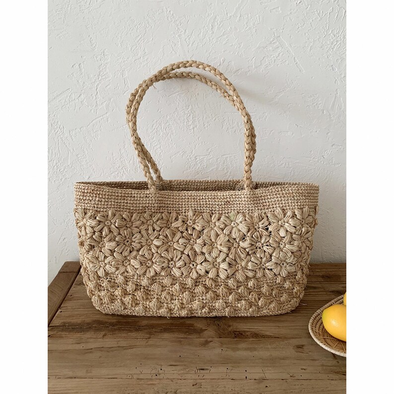 Op de afbeelding: Een beige geweven draagtas met bloemendetails. De tas heeft gevlochten handvatten en een rechthoekige vorm. De tas is gemaakt van natuurlijke vezels en staat op een houten oppervlak.