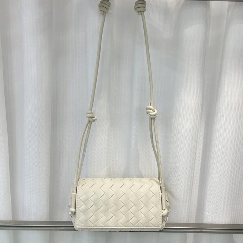 Op de afbeelding: Cr&egrave;mekleurige crossbodytas met een geweven patroon. De tas heeft een rechthoekige vorm met afgeronde hoeken en een lange, dunne riem. De riem is aan de boven- en onderkant geknoopt. De tas hangt voor een wit gordijn.