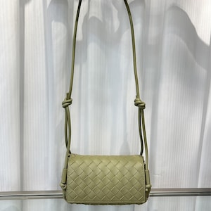 Op de afbeelding: Een lichtgroene crossbodytas met een geweven ontwerp. De tas heeft een lange, dunne riem met knopen aan de bovenkant. De tas is rechthoekig met een klepsluiting.