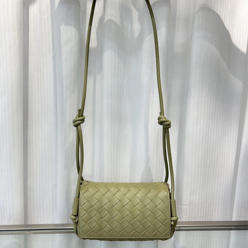 Op de afbeelding: Een lichtgroene crossbodytas met een geweven ontwerp. De tas heeft een lange, dunne riem met knopen aan de bovenkant. De tas is rechthoekig met een klepsluiting.