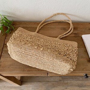 Op de afbeelding: Een beige geweven draagtas met gevlochten handvatten. De tas heeft een bloemenpatroon op het bovenste gedeelte en een rechthoekige vorm. De tas is gemaakt van een natuurlijk vezelmateriaal en ligt op een houten oppervlak.