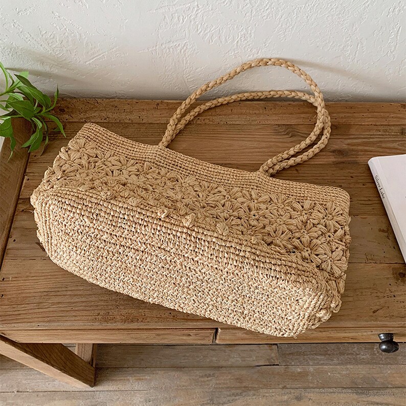 Op de afbeelding: Een beige geweven draagtas met gevlochten handvatten. De tas heeft een bloemenpatroon op het bovenste gedeelte en een rechthoekige vorm. De tas is gemaakt van een natuurlijk vezelmateriaal en ligt op een houten oppervlak.