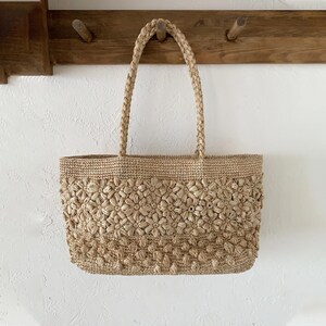 Op de afbeelding: Een beige geweven draagtas met een bloemenpatroon. De tas heeft gevlochten handvatten en hangt aan een houten rek. De body van de tas is rechthoekig met een gestructureerd, bloemachtig ontwerp.