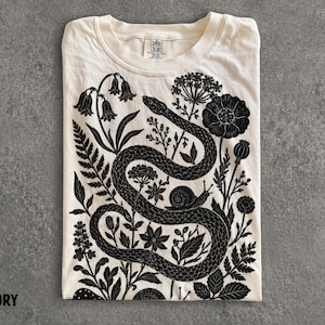 May include: An ivory t-shirt featuring a black graphic print of a coiled snake surrounded by various botanical elements including flowers, ferns, and a small snail. The shirt has a crew neckline and a Comfort Colors brand label at the inner neck.