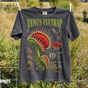 May include: A charcoal grey t-shirt featuring a detailed botanical illustration of a Venus flytrap. The graphic includes the text "VENUS FLYTRAP" and "DIONAEA MUSCIPULA" with various diagrams and labels explaining the plant's anatomy and trap mechanism.