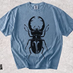 May include: A blue jean colored short-sleeve t-shirt featuring a large, detailed black graphic print of a stag beetle centered on the front. The shirt has a crew neckline and a visible brand label at the inner collar. The fabric has a washed texture.