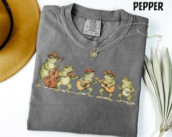 Puede incluir: Camiseta gris Comfort Colors con una ilustración de una banda de ranas tocando música. Las ranas llevan sombreros y tocan el violonchelo, el violín, guitarras y un banjo. La palabra "Pepper" está impresa en la parte superior.