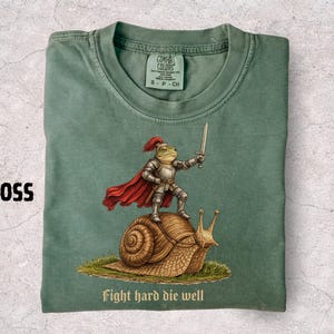 Puede incluir: Una camiseta de color verde musgo con un gráfico de una rana vestida con armadura de caballero plateada y capa roja, sosteniendo una espada mientras está de pie sobre un caracol marrón. El texto "Fight hard die well" aparece debajo.