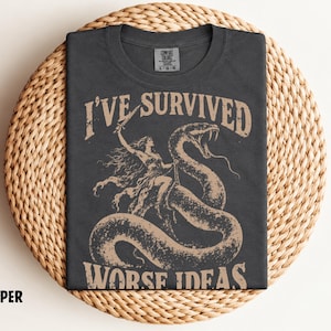 May include: A charcoal grey t-shirt featuring a tan graphic print of a figure holding a sword while riding a large snake. The text above the graphic reads I'VE SURVIVED and the text below reads WORSE IDEAS. The shirt has a Comfort Colors label.
