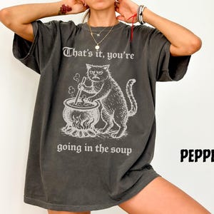 May include: Dark grey t-shirt featuring a grumpy cat stirring a cauldron with the text "That's it, you're going in the soup." The shirt has a vintage, distressed look and the word "PEPPER" is printed on the bottom right.