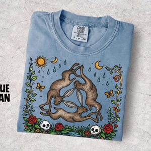 May include: Blue jean colored t-shirt with a round neck. The shirt features a graphic of three hares in a triangle, surrounded by floral and celestial elements. The text "BLUE JEAN" is on the left side of the shirt.