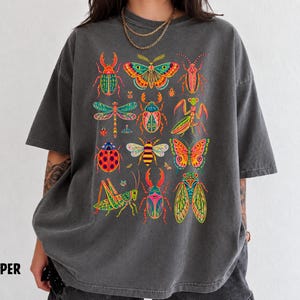 May include: A charcoal grey short-sleeve t-shirt featuring a vibrant, multi-coloured graphic print of various insects, including beetles, butterflies, a dragonfly, a praying mantis, a bee, and a cicada, arranged in a grid-like pattern on the front.