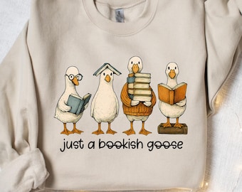 Può includere: Felpa beige con una stravagante illustrazione di quattro oche che leggono. Una indossa gli occhiali, un'altra ha un tetto a forma di libro. Il testo "just a bookish goose" è stampato sotto. Un design accogliente a tema letterario.