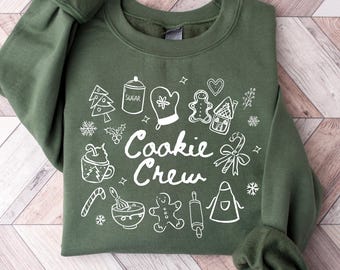 May include: An olive green sweatshirt with the words "Cookie Crew" in white script, surrounded by white illustrations of baking-related items like a gingerbread house, rolling pin, and oven mitt. The sweatshirt is made of a soft material.