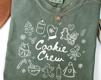 May include: Olive green t-shirt with a white graphic design featuring cookie-themed illustrations and the words "Cookie Crew." A gold necklace with a pendant rests on the shirt. The shirt is folded, with a brown hat and dried palm leaves in the background.