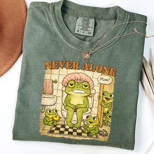 May include: A sage green Comfort Colors t-shirt with the words "NEVER ALONE" and a cartoon frog illustration. The frog is sitting on a toilet, wearing a shower cap, with other frogs nearby. A silver necklace is draped across the shirt.