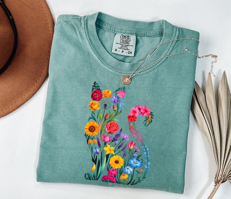 Op de afbeelding: Een zeegroen t-shirt met een bloemenkatontwerp. De kat is samengesteld uit kleurrijke bloemen, waaronder zonnebloemen, rozen en blauwe klokjes. Het shirt heeft een ronde hals en korte mouwen. Een zilveren ketting met een kleine hanger ligt over het shirt.
