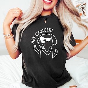 May include: Black t-shirt with the text "HEY CANCER!" and a white graphic of a dog wearing sunglasses and giving the middle finger. The shirt is a dark color and has a crew neck.