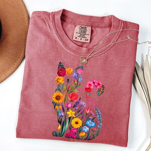 Op de afbeelding: Een stoffig roze Comfort Colors t-shirt met een bloemenkatontwerp. De kat is samengesteld uit kleurrijke bloemen, waaronder zonnebloemen, rozen en blauwe klokjes. Een zilveren ketting met een hanger rust op het shirt. Het shirt is maat S-P-CH.